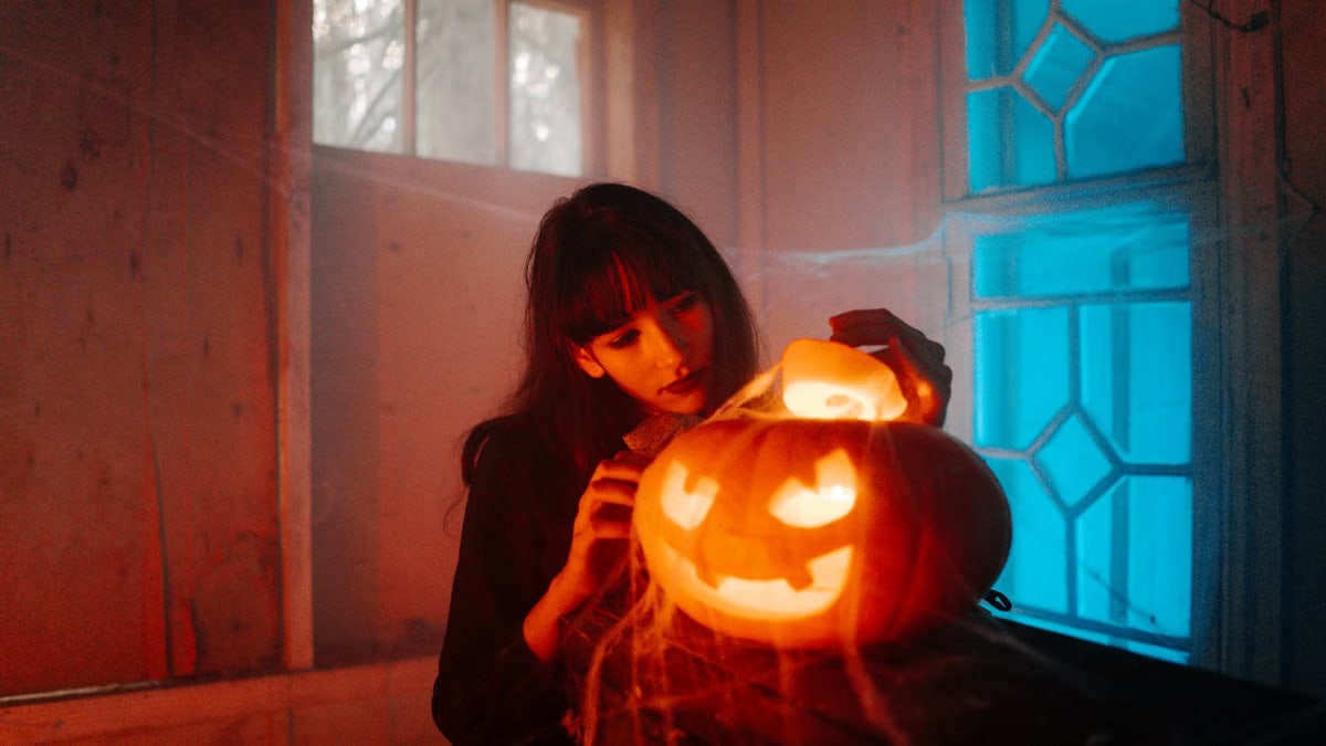 A woman carves a glowing jack-o'-lantern indoors amidst cobwebs and eerie lighting, celebrating Halloween.